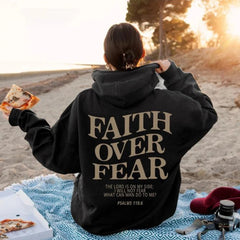 Woman wearing a Faith Over Fear hoodie on the beach, showcasing its inspirational message and cozy design.